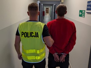 policjant trzyma zatrzymanego mężczyznę, obaj stoją tyłem, zatrzymany ma na rękach założone kajdanki