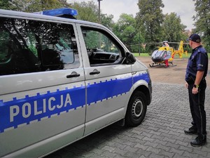 Zdjęcia kolorowe: Umundurowany policjant stojący obok oznakowanego radiowozu Policji.