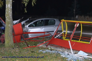 Widok miejsca wypadku. Przewrócony słup ogłoszeniowy, zniszczony przystanek i uszkodzony Peugeot 307 przed tablicą ogłoszeniową.