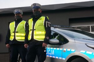 Na zdjęciu widoczni dwaj umundurowani policjanci, stojący na tle oznakowanego radiowozu.