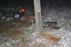 Na zdjęciu widoczny betonowy słup, obok którego na ziemi znajduje się kilka sztuk sanek. W tle częściowo widoczny ciągnik.