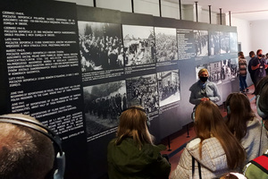 Wizyta w miejscu pamięci Auschwitz. Na zdjęciu widoczna młodzież słuchająca przewodniczki, za której plecami usytuowane są tablice z czarno białymi zdjęciami i opisami zdjęć.