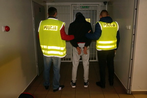 Na zdjęciu widoczni dwaj policjanci w kamizelkach odblaskowych z napisem POLICJA, którzy wyprowadzają z Pomieszczeń dla Osób Zatrzymanych, mężczyznę.