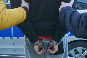 Na zdjęciu widoczny częściowo mężczyzna, który na ręce trzymane z tyłu ma założone kajdanki. Mężczyzna trzymany jest za ramiona przed dwóch policjantów. W tle widoczny radiowóz.