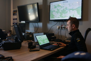 Na zdjęciu widoczny ratownik, który siedząc przed laptopem omawia działania ratownicze. W tle widoczny telewizor z wyświetloną na ekranie mapą.