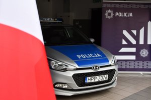Na zdjęciu widoczna częściowo polska flaga, radiowóz, a za nim policyjny baner z gwiazdą policyjna i napisem Policja.