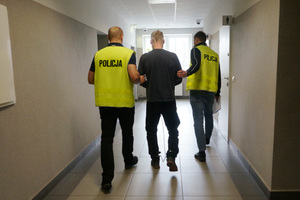 Na zdjęciu widoczni dwaj policjanci ubrani w kamizelki odblaskowe z napisem POLICJA, którzy prowadzą korytarzem sprawcę rozboju. Mężczyzna ma kajdanki zespolone założone na ręce i na nogi.