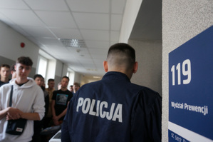 Policjant oprowadza uczniów po budynku komendy. Na ścianie widoczna tabliczka z napisem: Wydział Prewencji