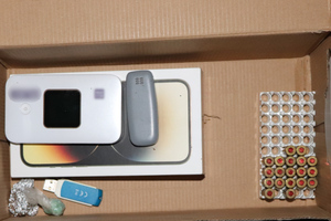 Tekturowe pudełko, w którym po prawej stronie widoczne są w tak zwanym koszyczku naboje, a po lewej stronie sprzęty elektroniczne wraz z foliowymi zwitkami.