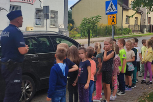 Umundurowany policjant prowadzi spotkanie z dziećmi przy ulicy, w rejonie oznakowanego przejścia dla pieszych.