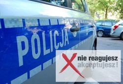 Częściowo widoczny radiowóz z napisem POLICJA na drzwiach. W prawym dolnym rogu napis: Nie reagujesz akceptujesz!