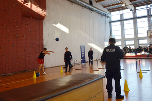 Na zdjęciu widoczny zawodnik, który rzuca piłką. Obok widoczni umundurowani policjanci.