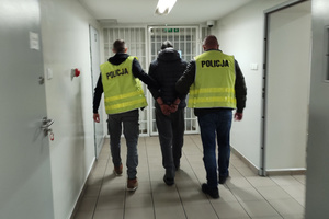 Na zdjęciu widoczni są dwaj nieumundurowani policjanci, w kamizelkach odblaskowych z napisem POLICJA, którzy prowadzą korytarzem zatrzymanego mężczyznę. Mężczyzna na ręce trzymane z tyłu ma założone kajdanki.