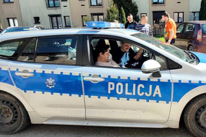 Zdjęcie: Warsztaty Terapii Zajęciowej w Łazach. Na zdjęciu widoczny jest umundurowany policjant, który siedzi za kierownicą radiowozu, a obok niego siedzi uczestniczka warsztatu.