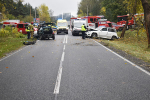 Na zdjęciu widoczne miejsce zdarzenia drogowego - dwa rozbite samochody, a także wozy strażackie i ambulans. Widoczni także policjanci, wykonujący na miejscu czynności.