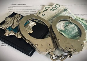 Zdjęcie pochodzi ze strony internetowej Policji Mazowieckiej. Na zdjęciu widoczne trzy banknoty 100-złotowe, a na nich położone kajdanki. W tle zapis artykułu 229 kodeksu karnego - przekupstwo.