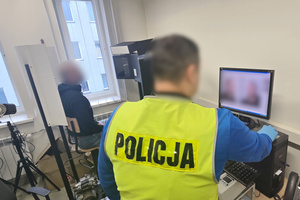 Na zdjęciu widoczny nieumundurowany policjant w kamizelce odblaskowej z napisem POLICJA, który wykonuje zdjęcia sygnalityczne zatrzymanemu mężczyźnie.