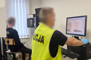 Na zdjęciu widoczny technik kryminalistyki i zatrzymany mężczyzna, któremu robi zdjęcie sygnalityczne.