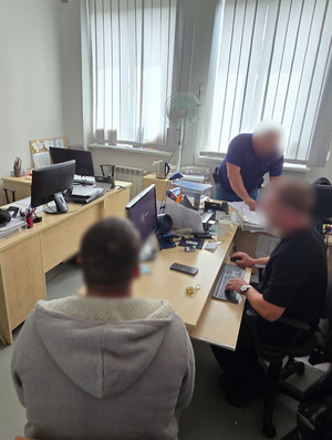 Na zdjęciu widoczne pomieszczenie biurowe oraz dwóch nieumundurowanych policjantów. Widoczny jest także zatrzymany mężczyzna, który siedzi przy biurku, przy którym prowadzi czynności jeden z policjantów.
