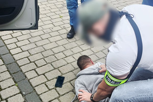 Na zdjęciu widoczni nieumundurowany policjant, który przytrzymuje leżącego na ziemi zatrzymanego mężczyznę.