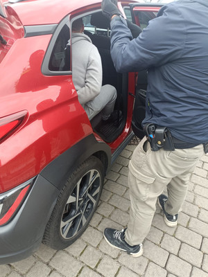 Na zdjęciu widoczny nieumundurowany policjant stojący przy samochodzie, wewnątrz którego siedzi zatrzymany mężczyzna.