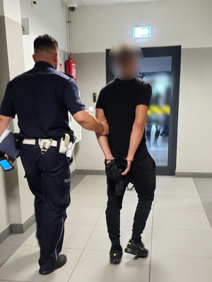 Na zdjęciu widoczny policjant, który prowadzi zatrzymanego mężczyznę. Mężczyzna ma założone kajdanki na ręce trzymane z tyłu.