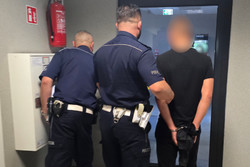 Na zdjęciu widoczni policjanci, którzy prowadzą zatrzymanego mężczyznę. Mężczyzna ma założone kajdanki na ręce trzymane z tyłu.
