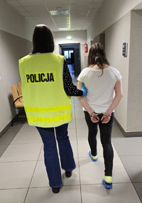 Na zdjęciu widoczna nieumundurowana policjantka w kamizelce odblaskowej z napisem Policja, która prowadzi korytarzem zatrzymaną kobietę. Zatrzymana ma założone kajdanki na ręce trzymane z tyłu.