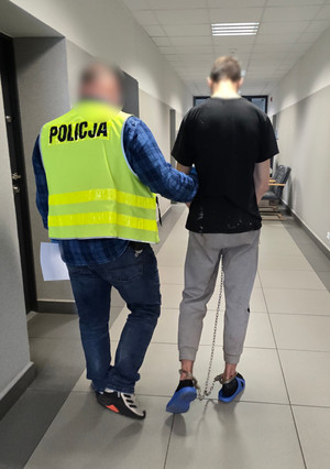 Na zdjęciu widoczny nieumundurowany policjant, który ma na sobie kamizelkę odblaskową z napisem: Policja. Policjant prowadzi zatrzymanego mężczyznę, który ma założone kajdanki zespolone.