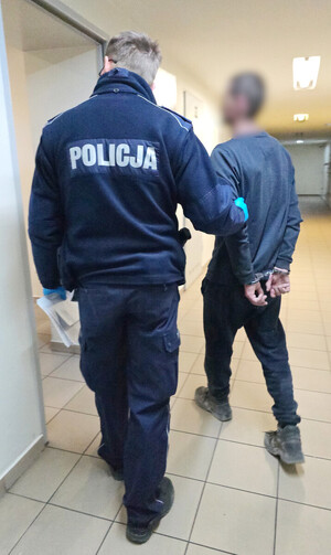 Na zdjęciu widoczny umundurowany policjant, który prowadzi zatrzymanego mężczyznę. Mężczyzna na rękach trzymanych z tyłu ma założone kajdanki.