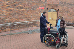 Na zdjęciu widoczny dzielnicowy oraz przedstawicielka Miejskiego Ośrodka Kultury w Zawierciu, której policjant przekazuje dwa wózki inwalidzkie oraz komplet kuli ortopedycznych.