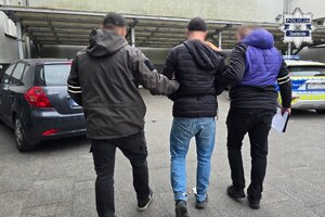 Nieumundurowani policjanci prowadzą do radiowozu zatrzymanego mężczyznę. Mężczyzna ma założone kajdanki zespolone.