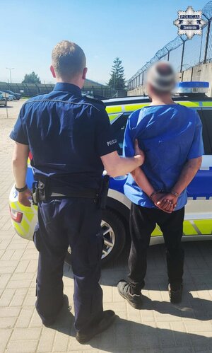 Policjant trzymający za ramię mężczyznę. Mężczyzna ma założone kajdanki na ręce trzymane z tyłu. Obydwaj stoją przy radiowozie.