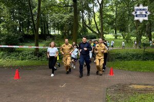 Policjant, nauczycielka, dwóch strażaków i dzieci biegną przez park.