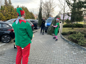 Na zdjęciu widać policyjny radiowóz, strażaka oraz mężczyznę przebranego za elfa trzymającego w rękach choinkę.