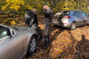 Na zdjęciu widać dwa szare pojazdy oraz policjanta z zatrzymanym mężczyzną stojących przy samochodzie.