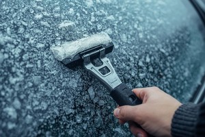 Grafika przedstawia szybę samochodową, która jest pokryta lodem i dłoń, która trzyma skrobaczkę do szyb.