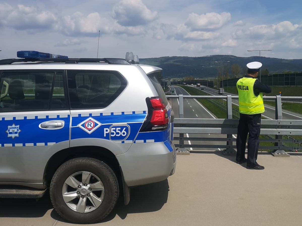 zdjęcie zrobione na wiadukcie po lewejstronie fragment radiowozu obok stoi policjant który dokonuje pomiaru w tle ulica i krajobraz