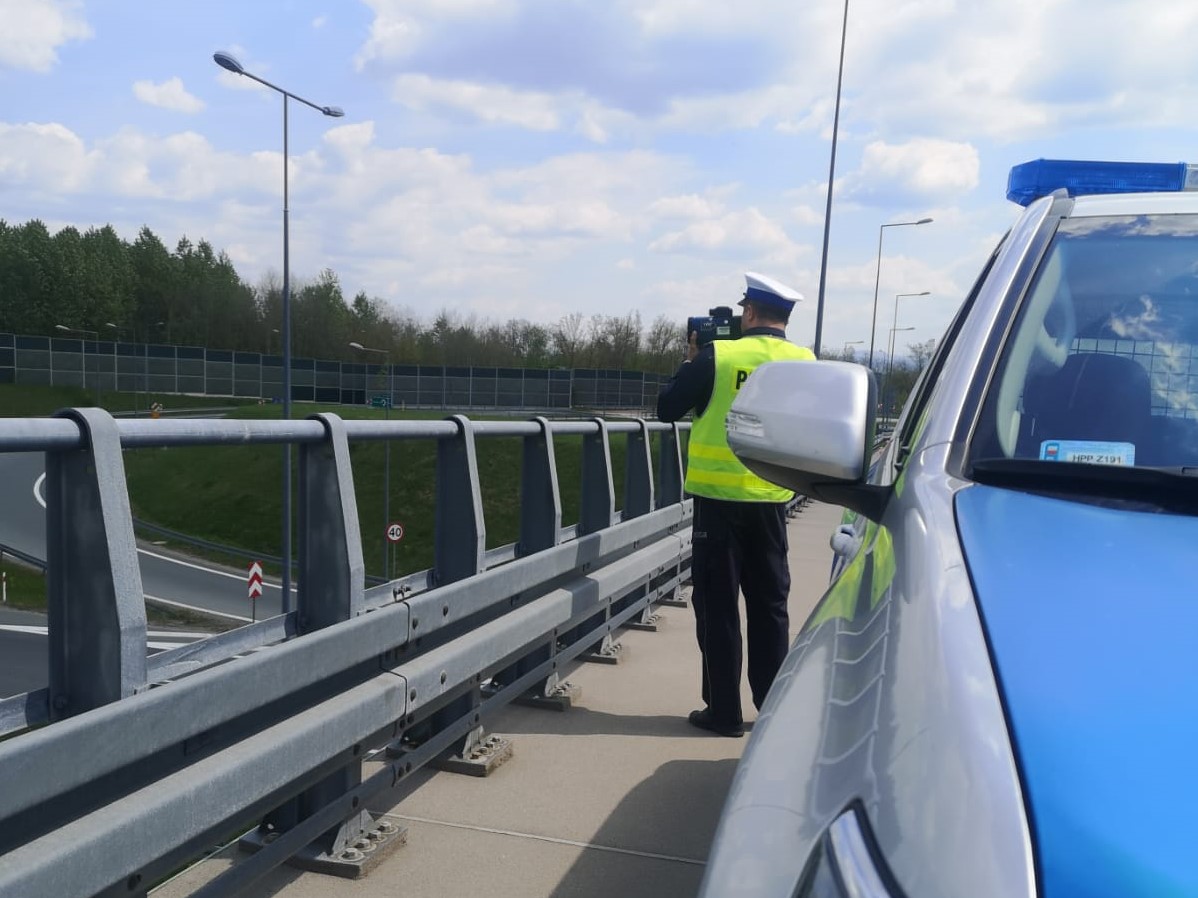 na pierwszym planie fragment radiowozu obok niego stoi policjant i dokonuje pomiaru w tle ulica