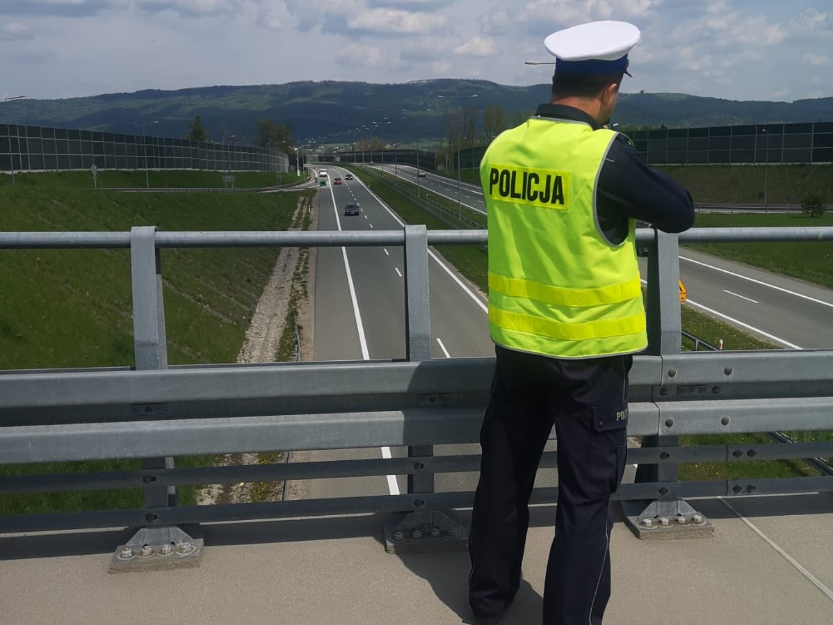 policjant  stoi tyłem na wiadukcie i dokonuje pomiaru w tle ulica samochody i góry