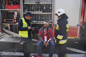 poszkodowany siedzi w pobliżu wozu strażackiego obok stoi policjant oraz strażak