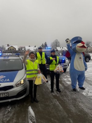 stoją policjantka pracownik word policjant i sznupek na tle radiwozów i machają dzieciom