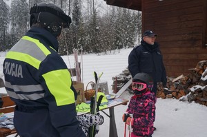 na zdjęciu kolorowym zrobionym na zewnątrz stoi bokiem policjant narciarz przed nim stoi dziewczynka