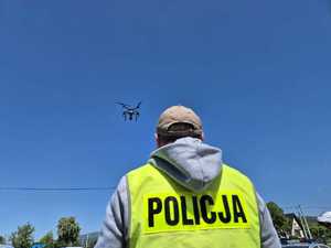 Zdjęcie kolorowe: fragment sylwetki policjanta w kamizelce z napisem Policja, który stoi tyłem - w tle widać latający dron