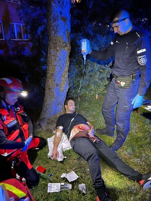zdjęcie kolorowe zrobione w nocy na ziemi leży poszkodowany z raną brzucha obok stoi policjant i trzyma kroplówkę