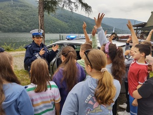 zdjęcie kolorowe dzieci stoją tyłem przed nimi policjantka prowadzi prelekcję, dzieci zgłaszają się, obok radiowóz w tle jezioro