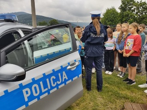 zdjęcie kolorowe na pierwszym planie radiowóz w tle policjantka stoi tyłem i prowadzi prelekcję przed nią stoją dzieci