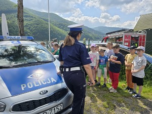 policjantka stoi tyłem prowadzi prelekcję dzieci stoją przed nią w tle jezioro