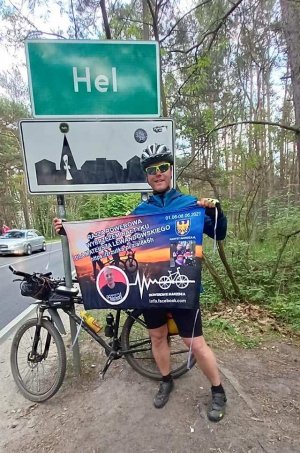Sierżant sztabowy Damian Lorisz z plakatem promującym przedsięwzięcie na tle znaku drogowego z nazwą miejscowości Hel.