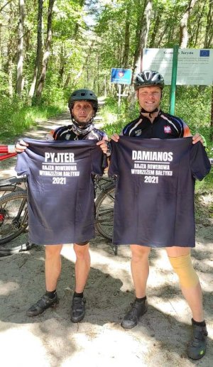 Sierżant sztabowy Damian Lorisz z towarzyszem wyprawy Piotrem w strojach rowerowych, trzymający okolicznościowe koszulki z nazwą przedsięwzięcia, na granicy polsko-rosyjskiej.